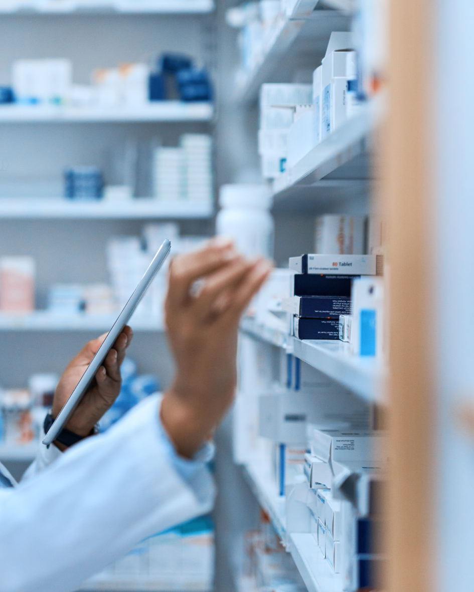 A pharmacist holding a tablet while checking medications on a shelf in a pharmacy.