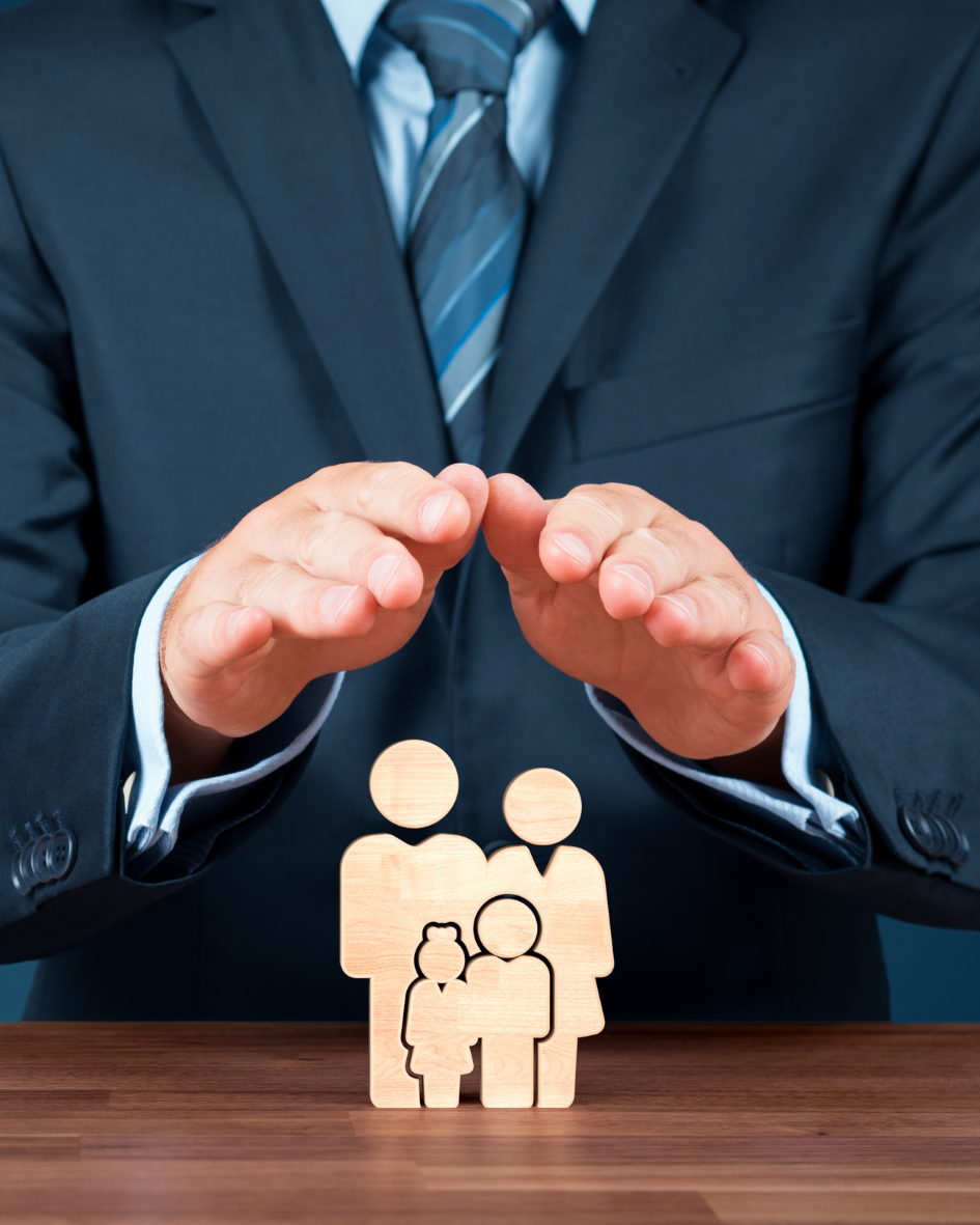 A businessman in a suit is placing his hands protectively over wooden figurines representing a family on a table.